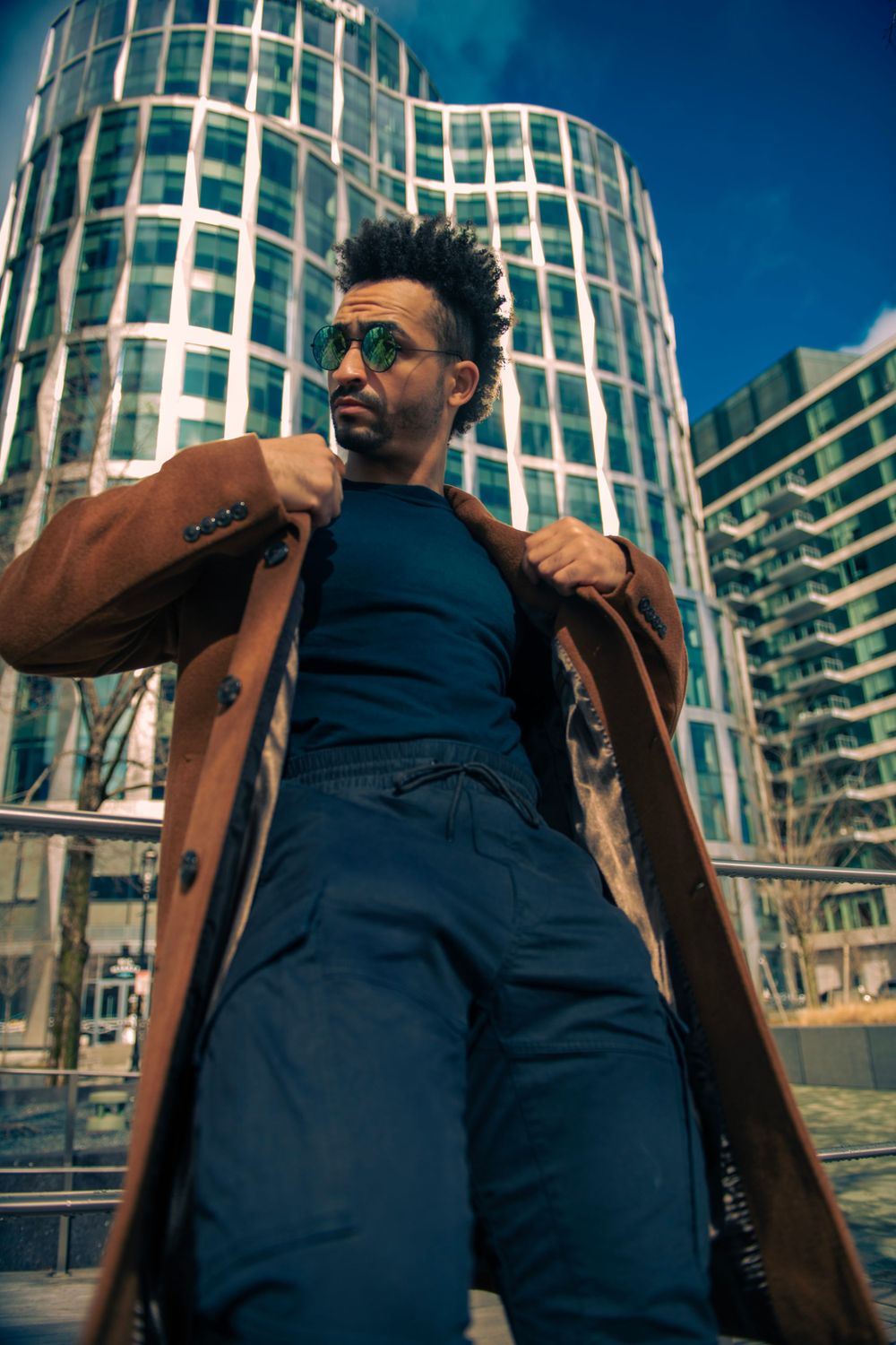 Urban fashion shot with person in camel coat and navy outfit against modern glass building.