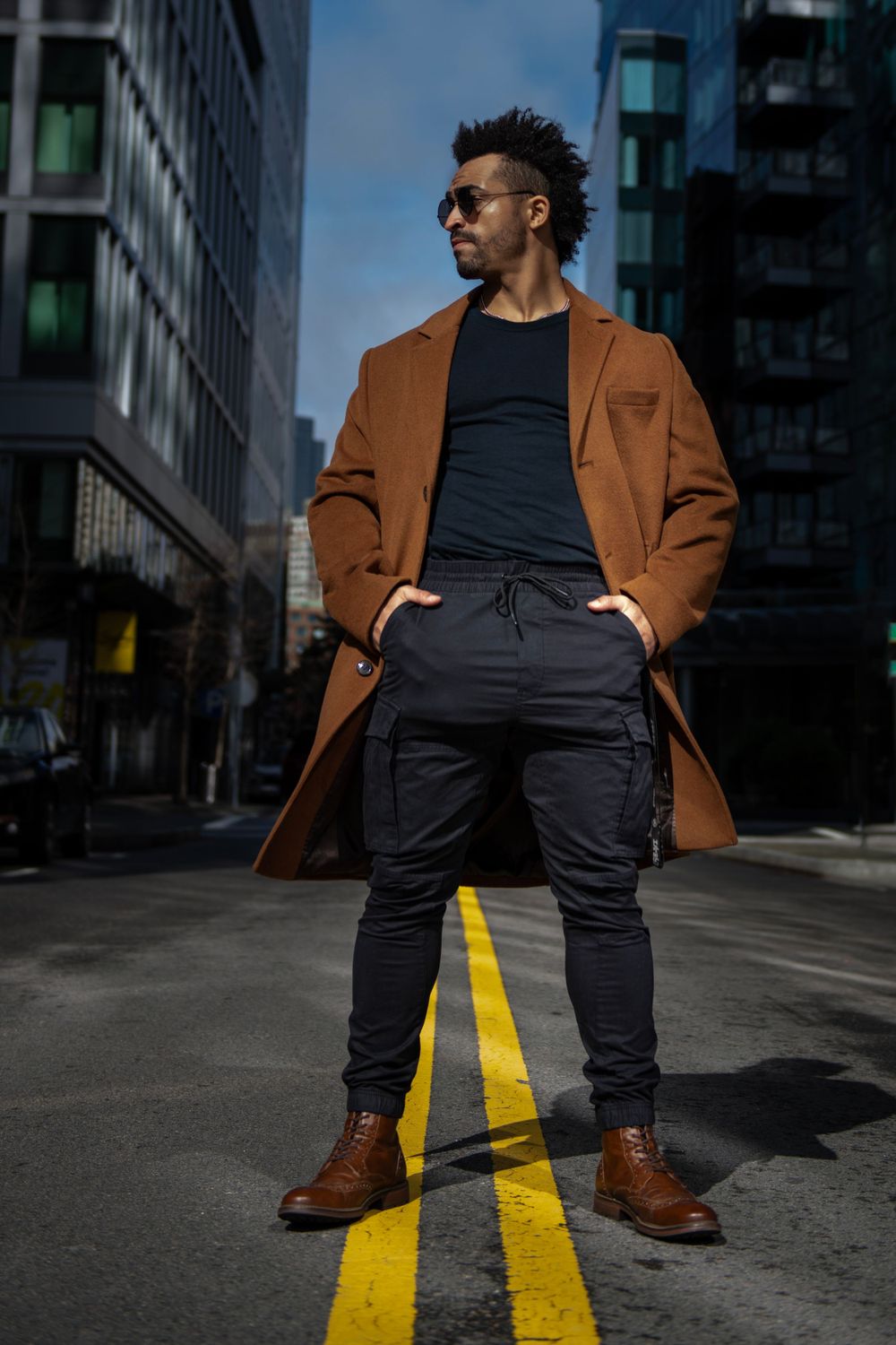 Street style photo of person in brown coat standing on yellow road line with buildings behind.