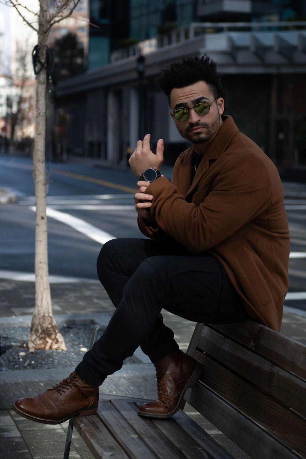 Fashion model in brown coat and boots sitting on bench in urban setting.