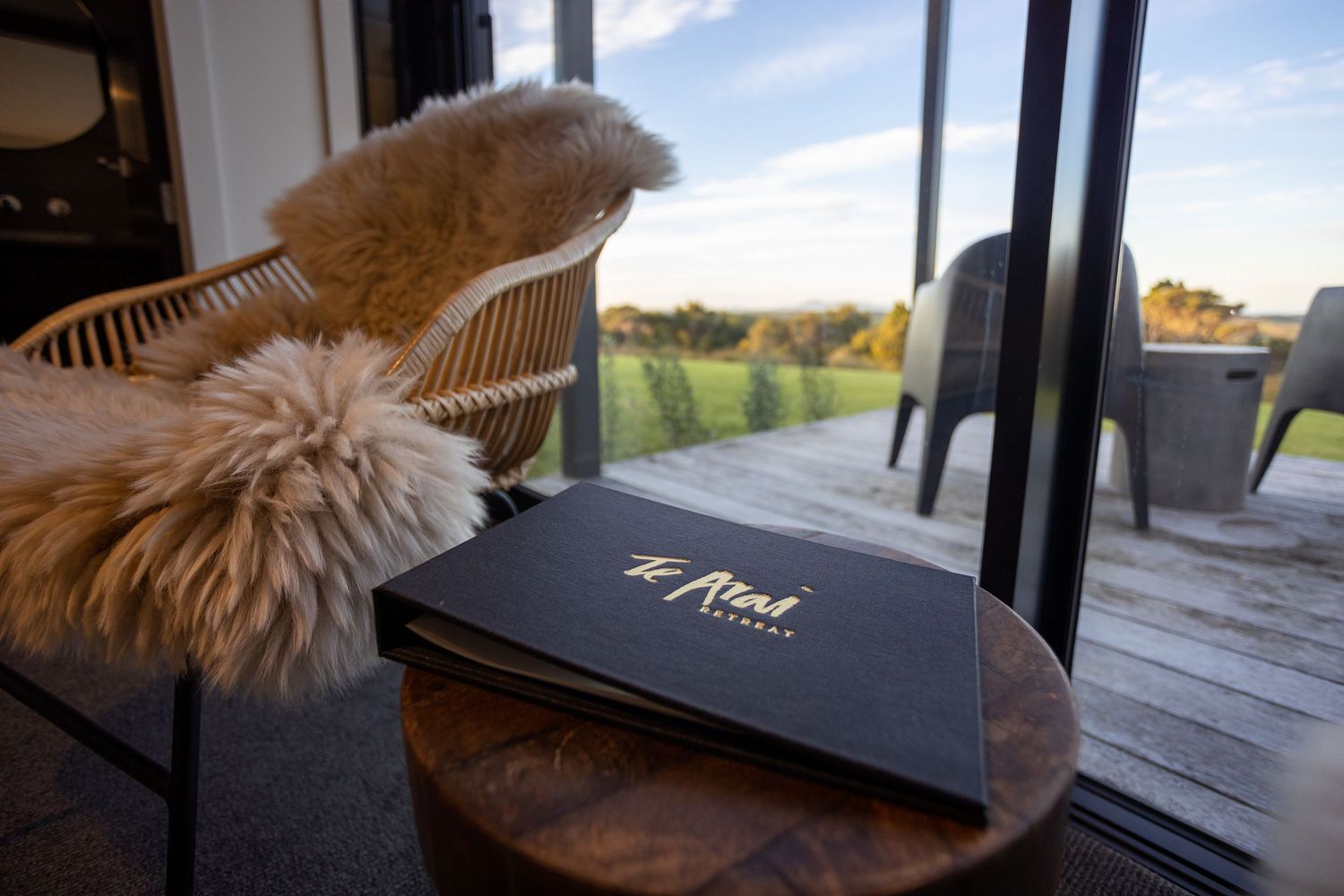 Luxury lodge menu book on wooden table beside sheepskin chair with scenic outdoor view.