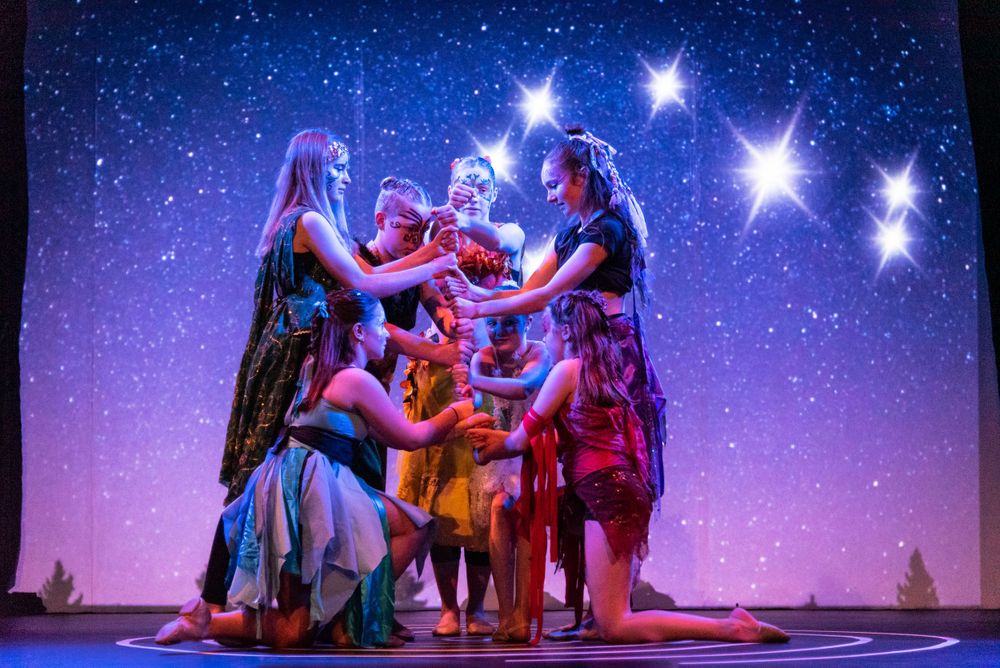 Performers in colorful costumes create a dramatic group pose against a starry purple backdrop.
