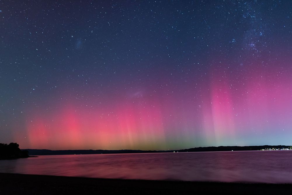 Vibrant pink and purple aurora borealis reflects over calm water against a starry night sky near a dark shoreline.