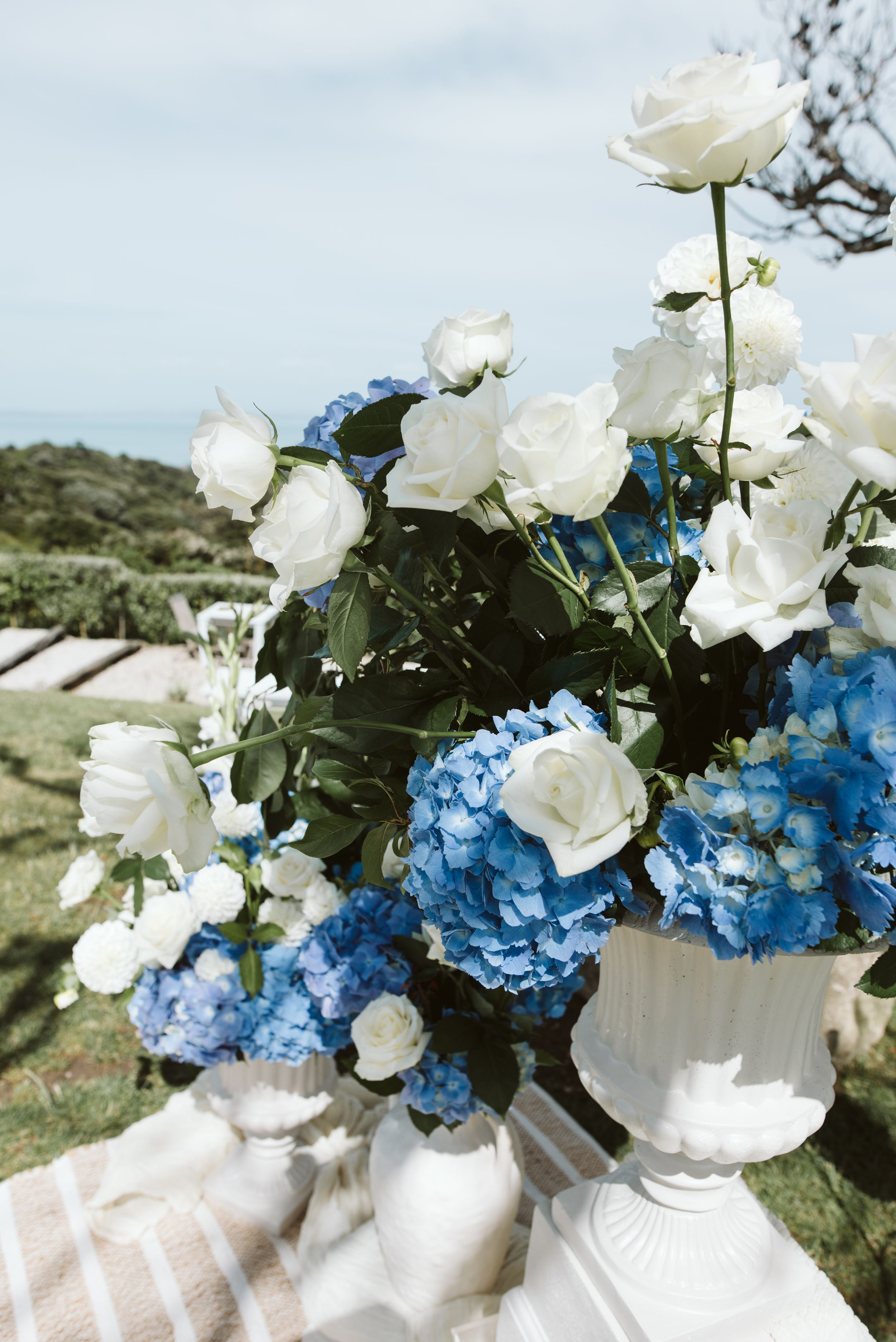 White roses and blue hydrangeas arranged in a decorative white vase against a coastal landscape backdrop.
