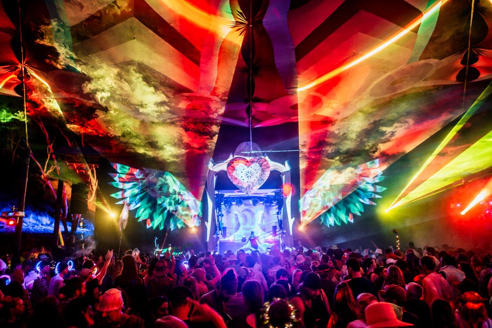 Colorful EDM festival with laser lights and angel wing visuals above dancing crowd.