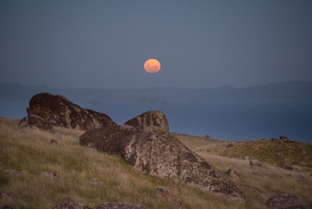A full orange moon rises above rocky terrain and distant mountains at dusk.