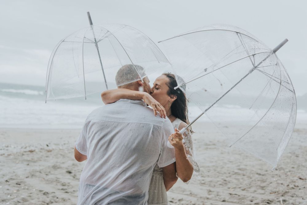 Couple sharing romantic moment under clear umbrellas on cloudy beach.