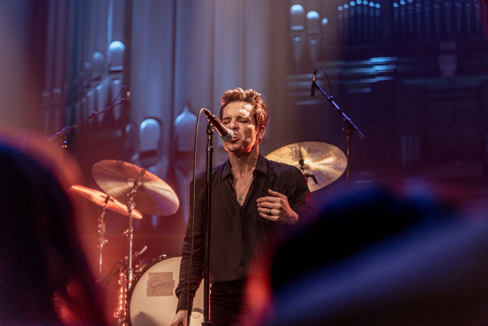 Concert performer singing on stage with dramatic lighting and drum kit in background.