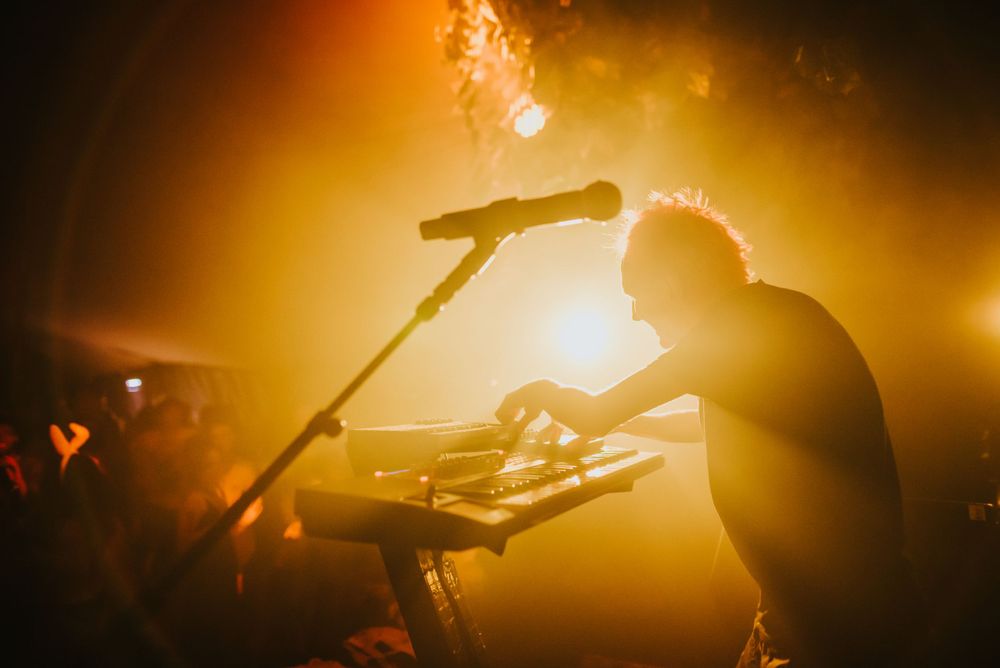 A silhouetted musician performs with equipment backlit by golden stage lights and smoke.