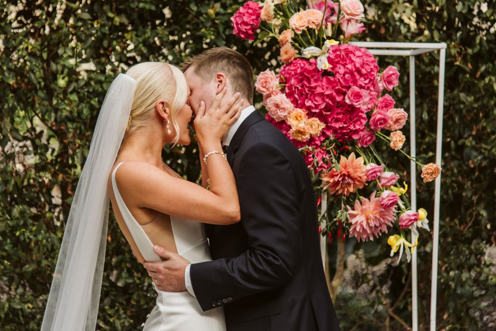 A romantic wedding kiss captured beside vibrant pink floral arrangements and greenery.