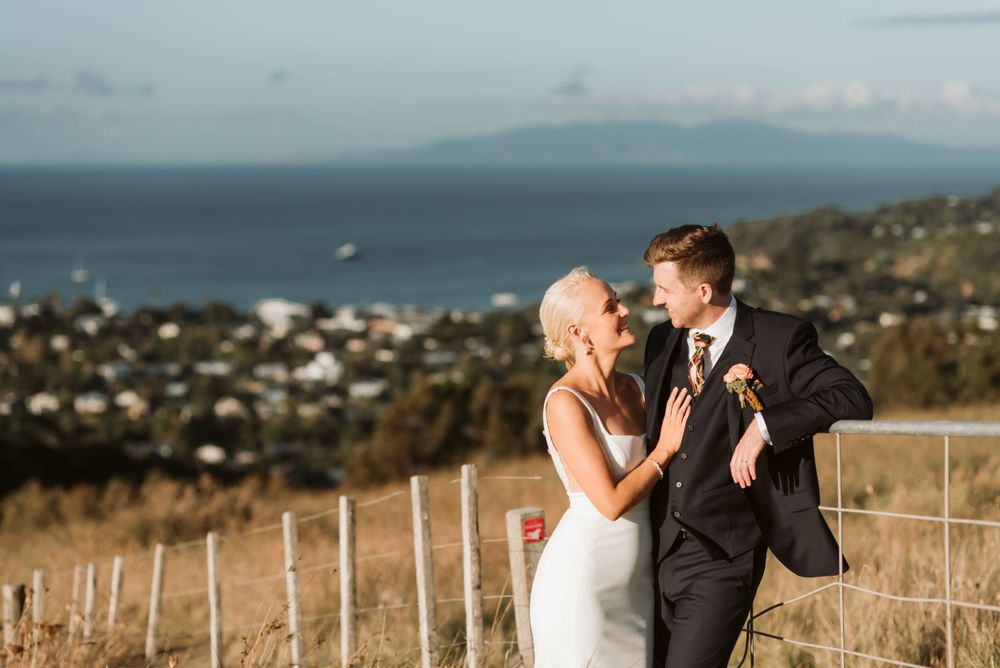 A couple in wedding attire embraces on a hilltop overlooking a coastal town and ocean vista during golden hour.
