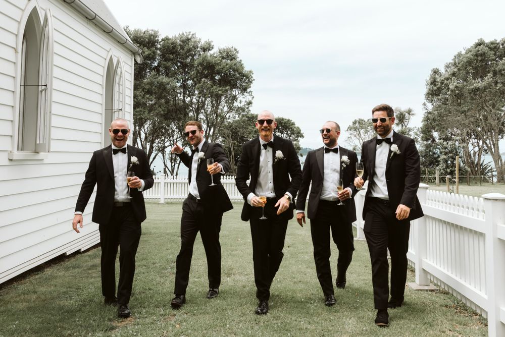 Groomsmen in black tuxedos walk together laughing along white picket fence at outdoor wedding venue.