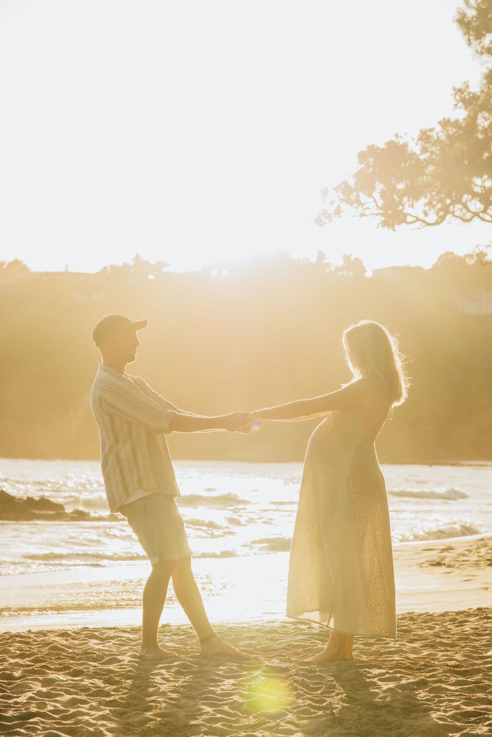 A romantic silhouette of two people holding hands on a beach at golden hour with sun flare in background.