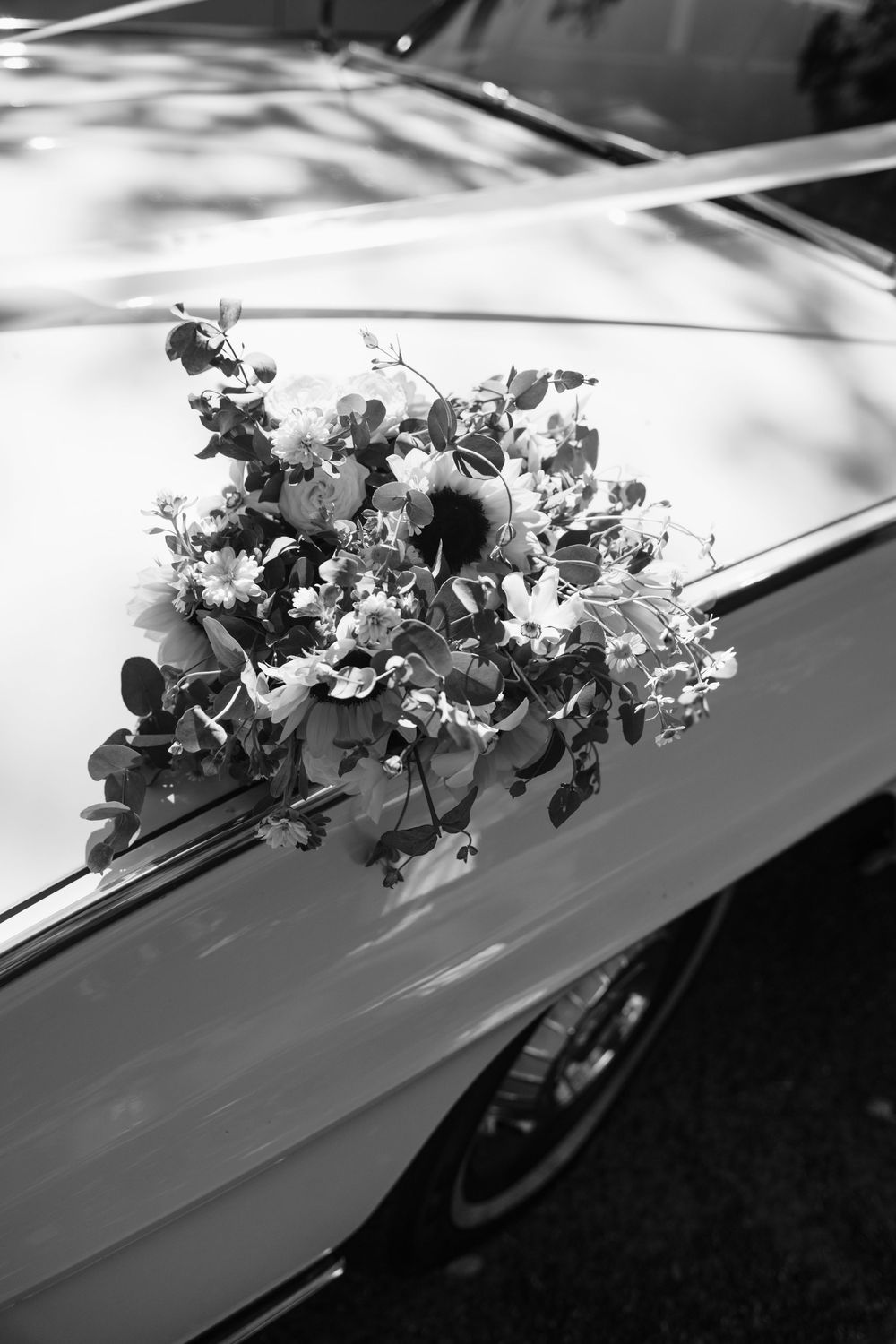 Black and white photo of floral arrangements adorning the hood of a classic car at a wedding ceremony.