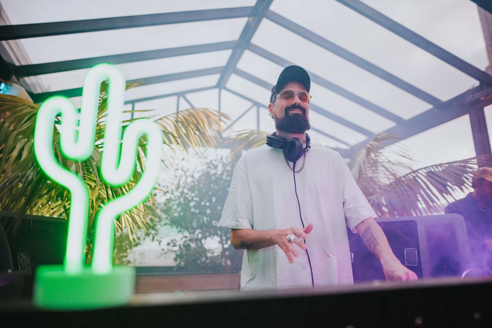 DJ performing at an outdoor venue with a neon green cactus sign illuminated in the background.