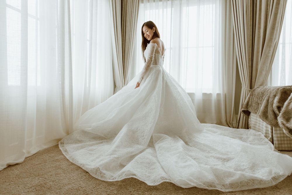 A flowing white wedding gown with long train swirls elegantly around a figure against light-filled windows.