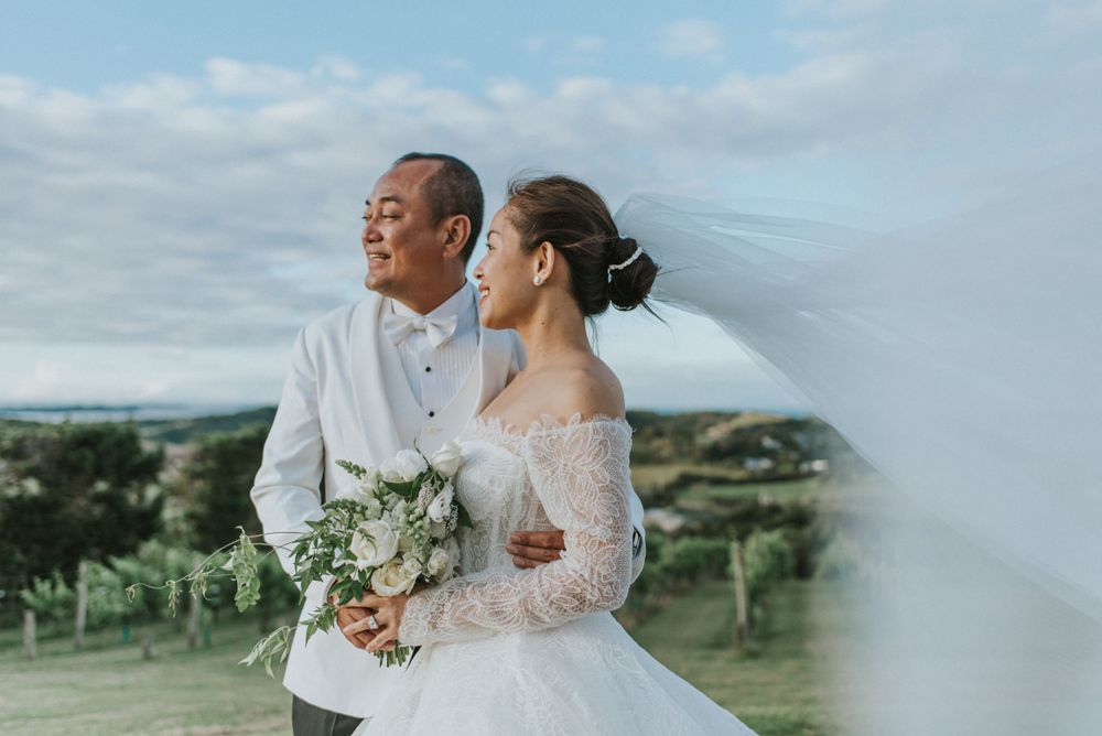 Newlyweds posing together on hilltop with flowing veil and bouquet.
