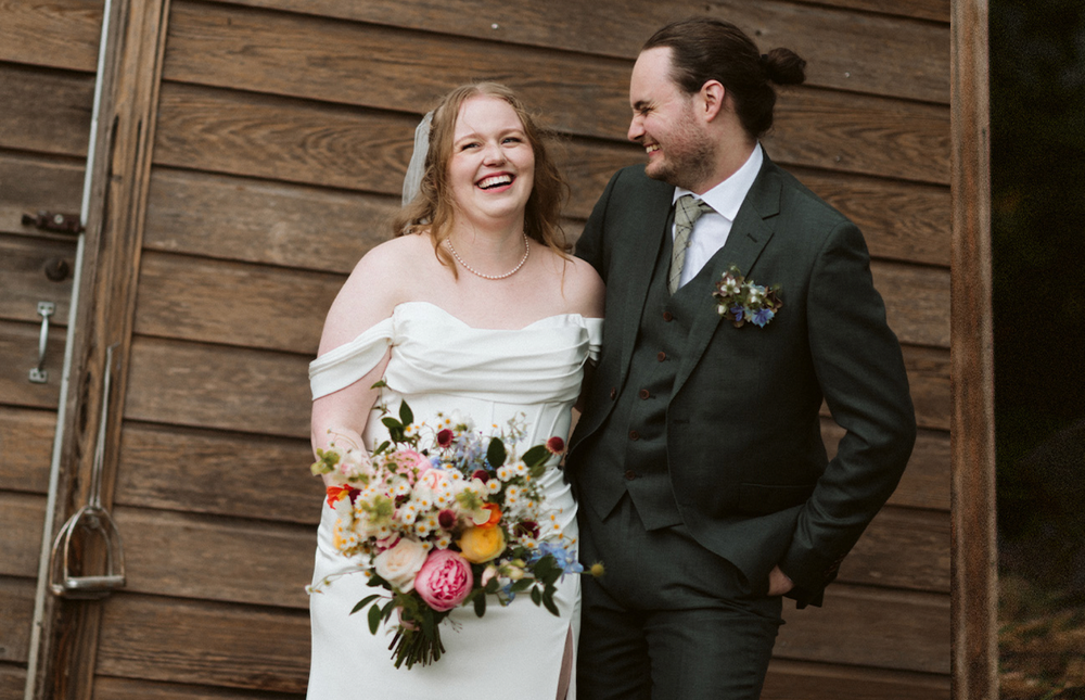 A couple shares a joyful moment in front of a rustic wooden wall with colorful wedding flowers.
