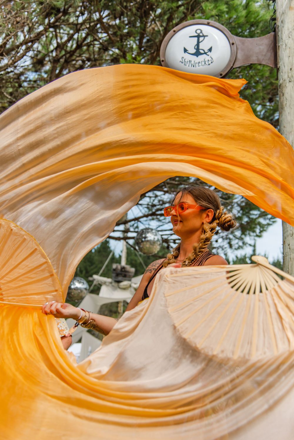 Dancer with flowing orange fabric performs beneath a vintage anchor sign outdoors.