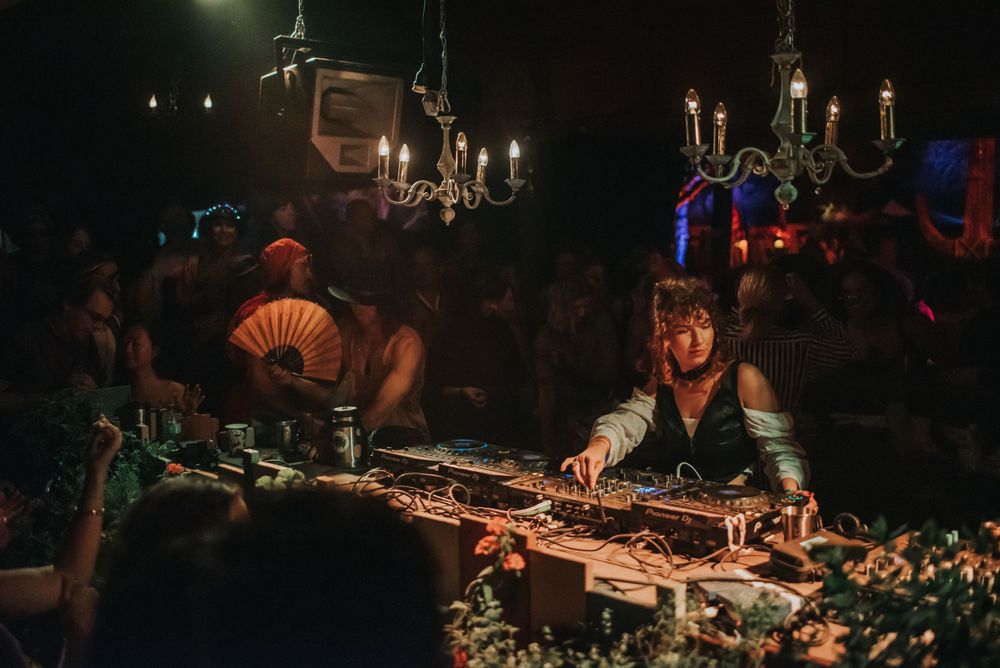 DJ performs at night behind equipment with ornate chandeliers and mood lighting overhead.