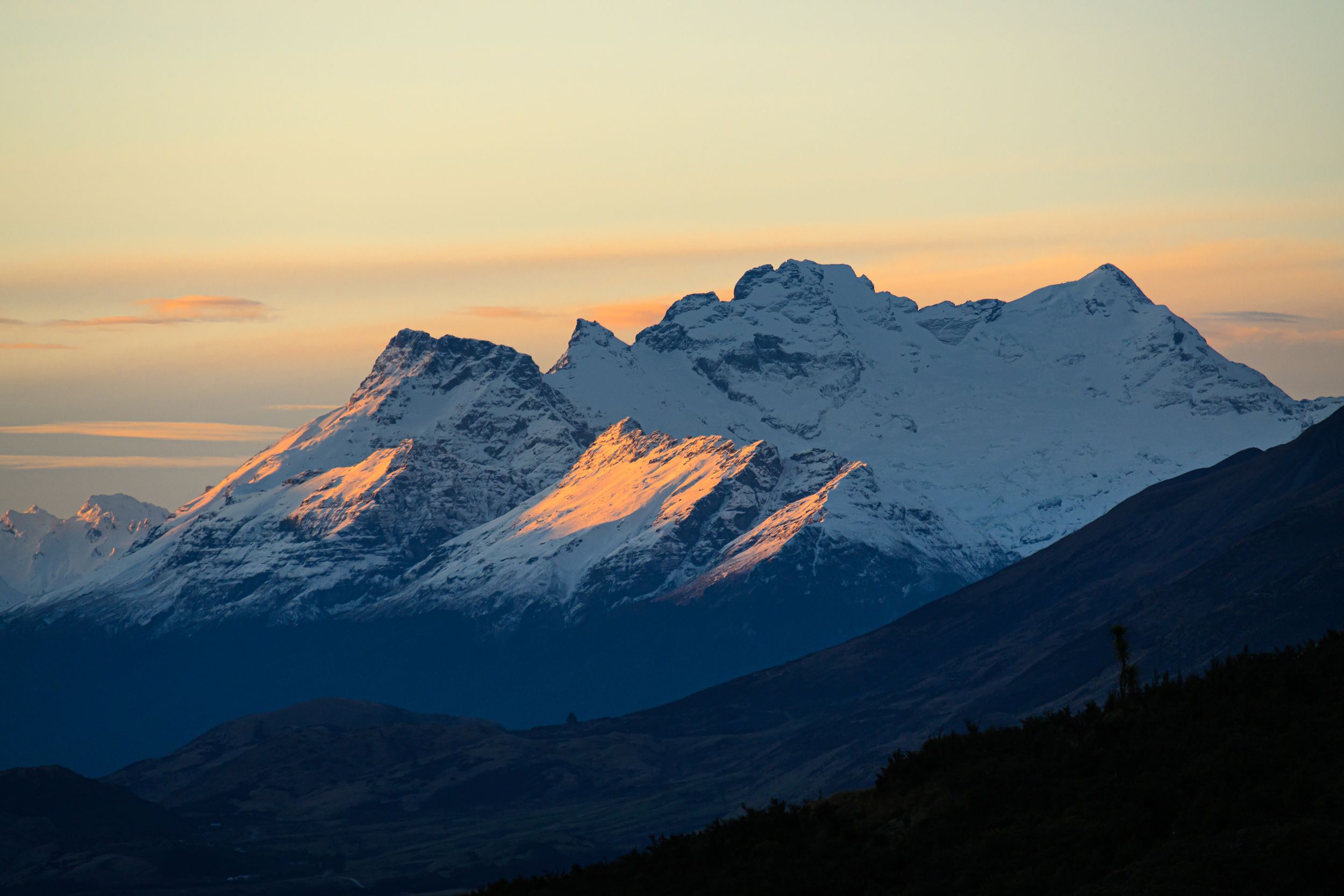Snow-capped mountain peaks reflect golden light at sunset against a darkening blue sky in a panoramic alpine landscape.