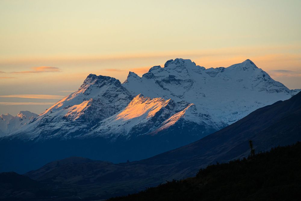 Snow-capped mountain peaks reflect golden light at sunset against a darkening blue sky in a panoramic alpine landscape.