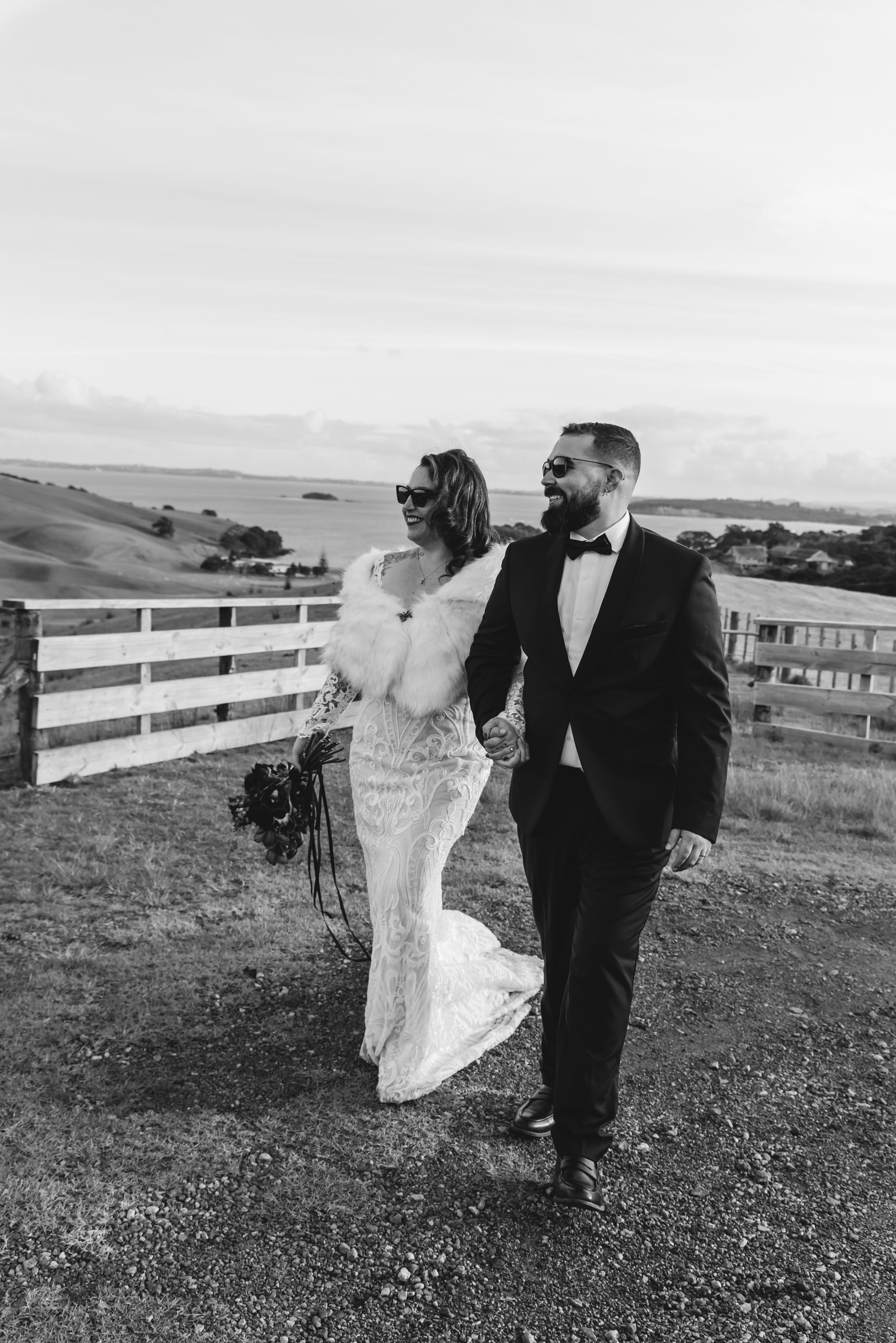 A couple shares a romantic moment in front of a white fence on rolling hills in black and white photography.