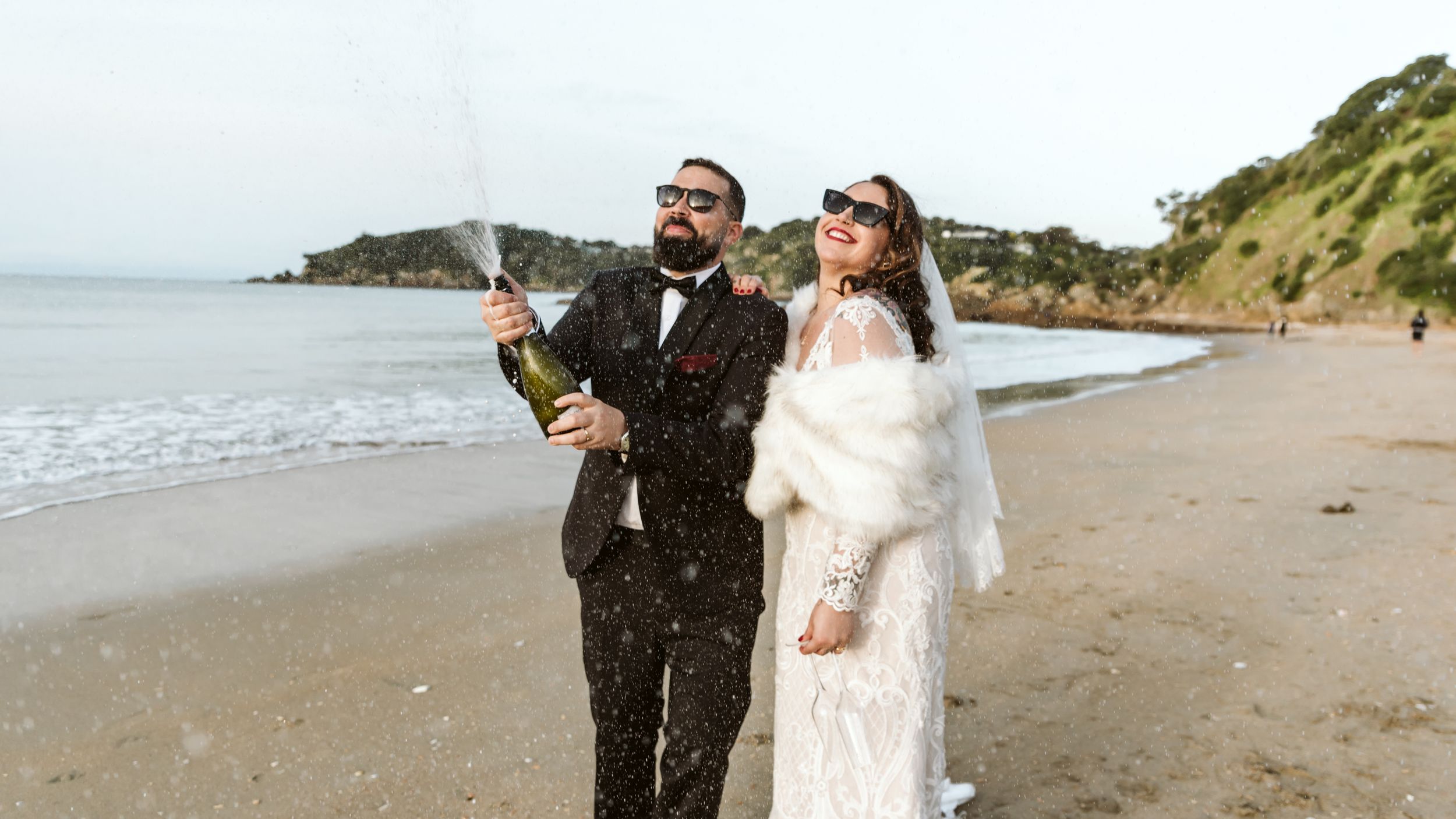 A couple celebrating with champagne on a scenic beach wearing formal wedding attire while waves crash behind them.