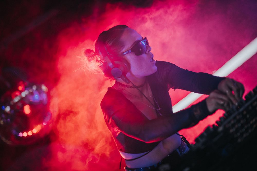 A DJ performs in red atmospheric smoke while working mixing equipment in a nightclub setting.