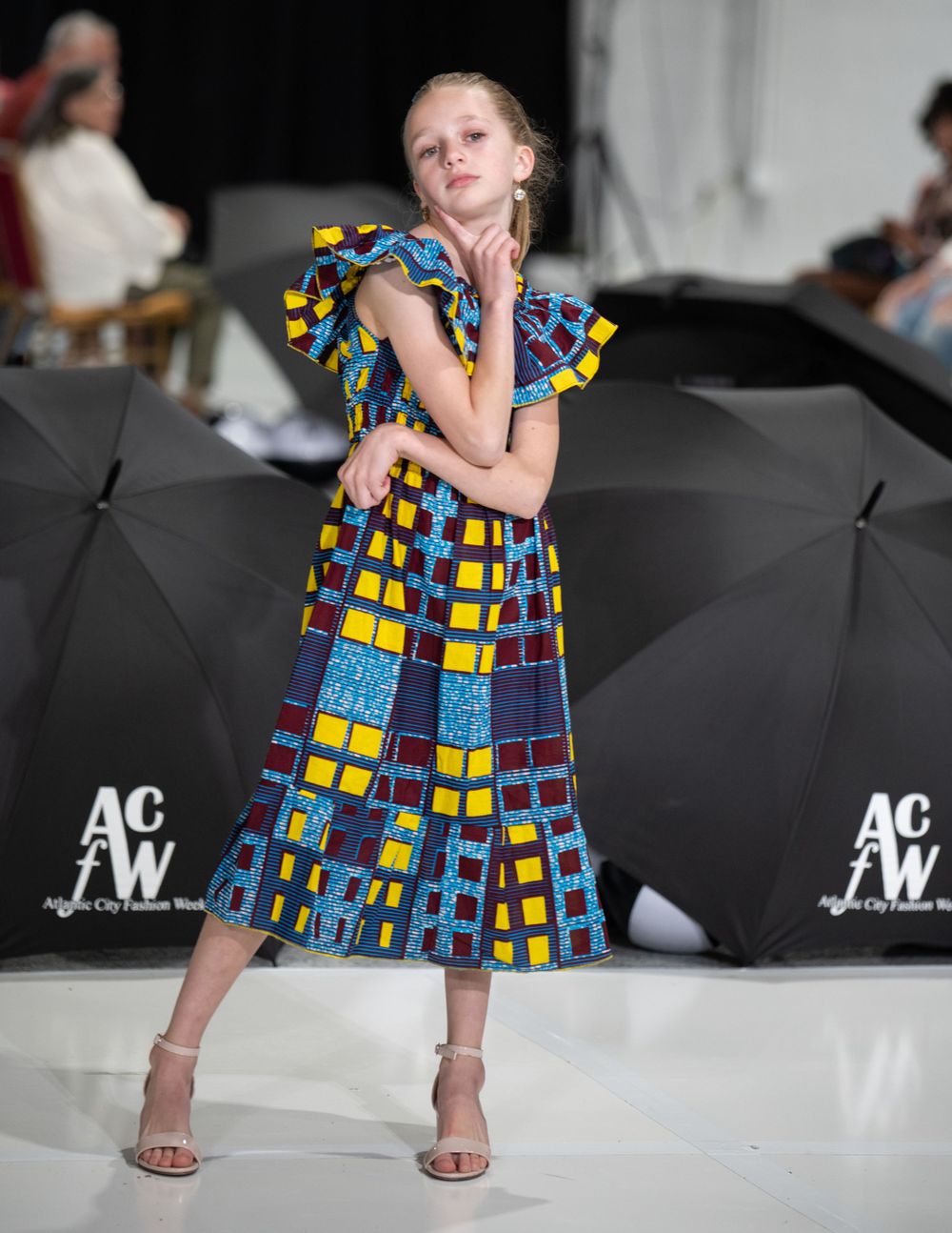 Young model walks runway in colorful geometric print dress with ruffled sleeves at fashion show.
