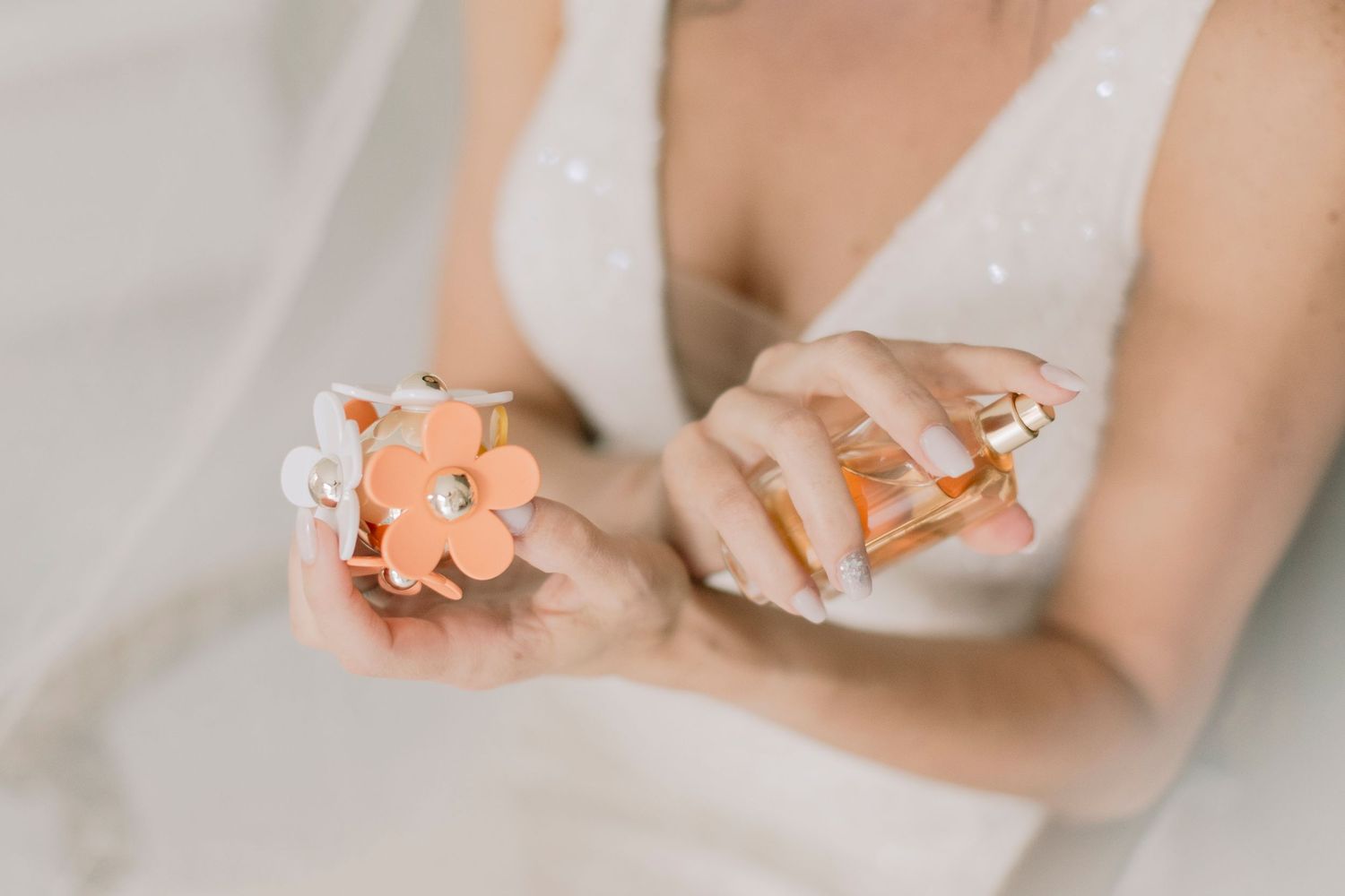Close-up of a bride holding an orange floral perfume bottle while wearing a sparkling white wedding dress.