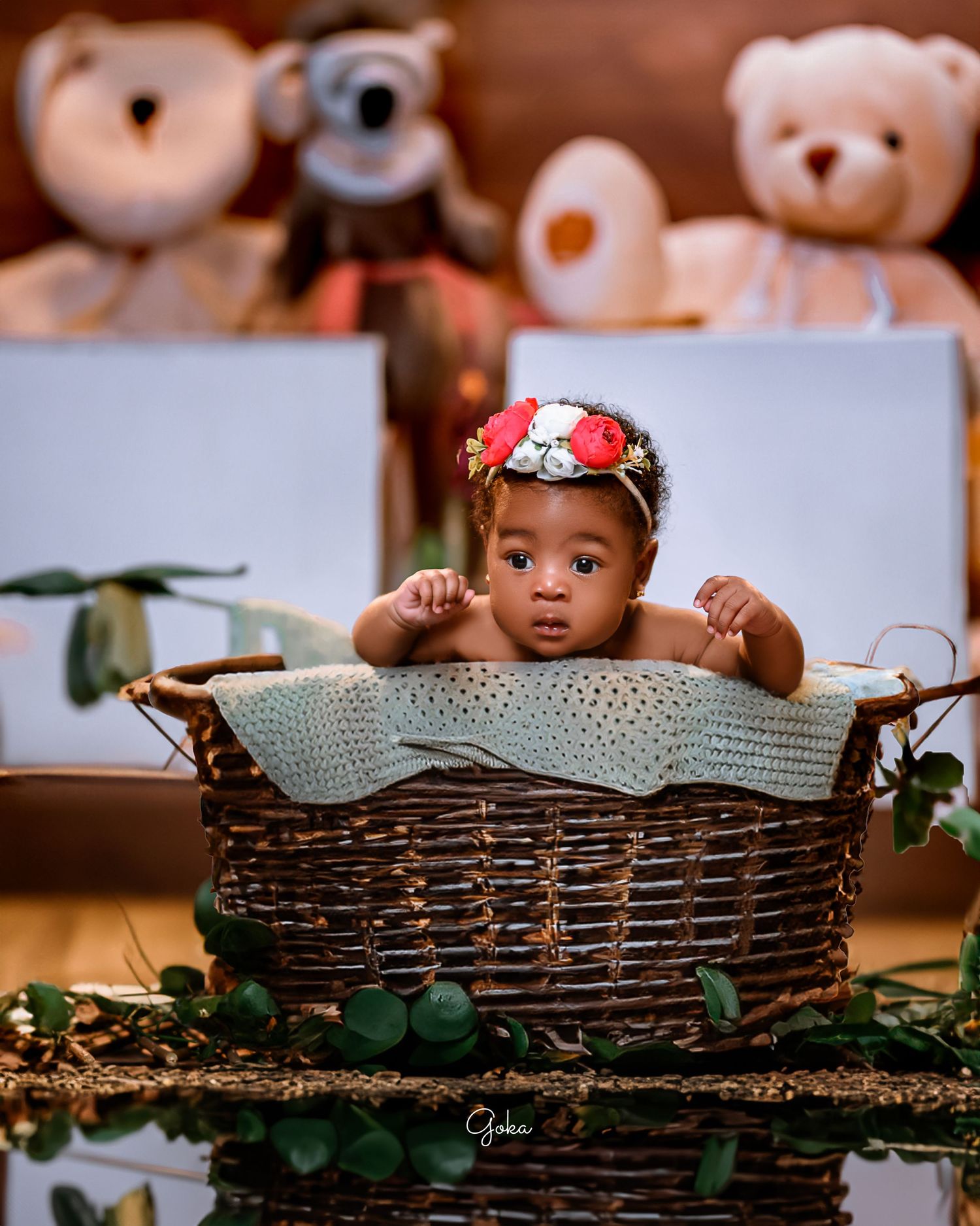 Baby lying in woven basket with floral headband surrounded by stuffed animals in studio setup.