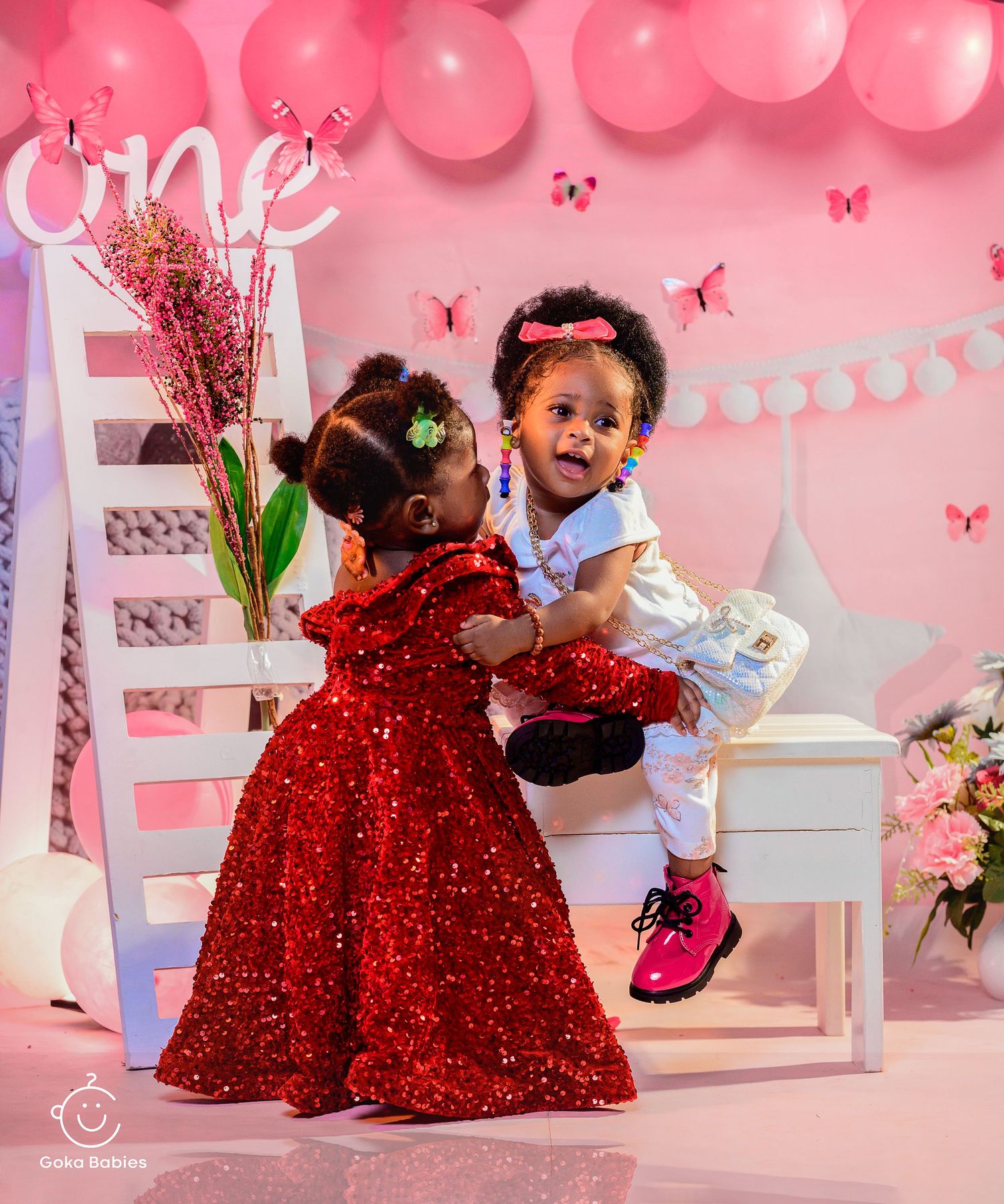 Two children in festive attire playing on white ladder against pink balloon backdrop.