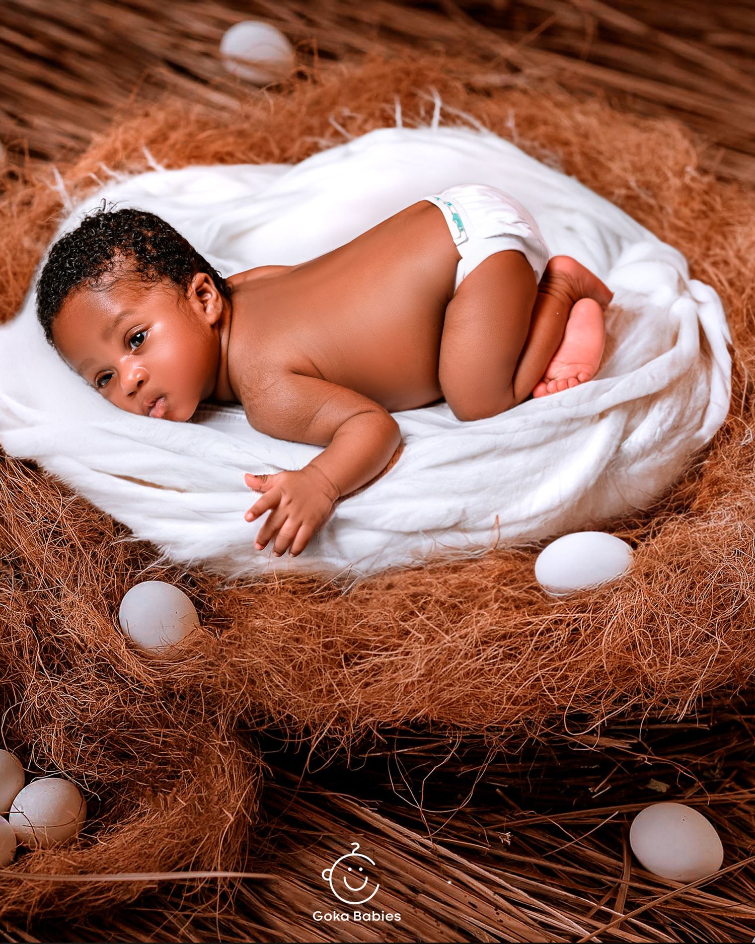 A newborn baby lying on soft white fabric surrounded by eggs and brown textured material in a studio setting.