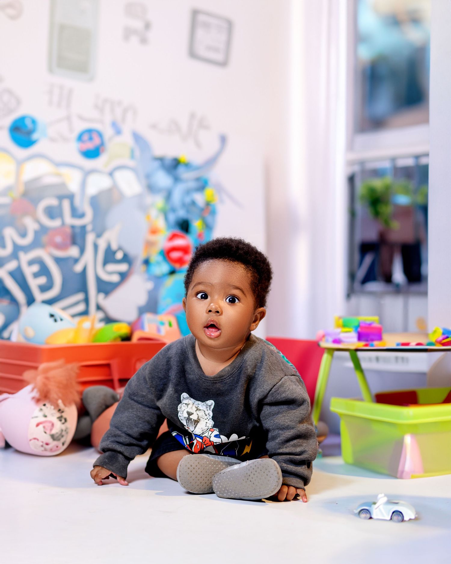 Baby in gray sweater playing with toys in colorful playroom setting.