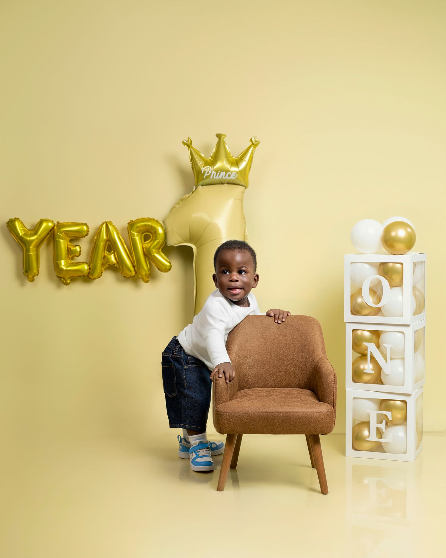 First birthday celebration setup with gold balloon letters spelling YEAR, a crown, brown chair, and white ONE blocks against yellow wall.