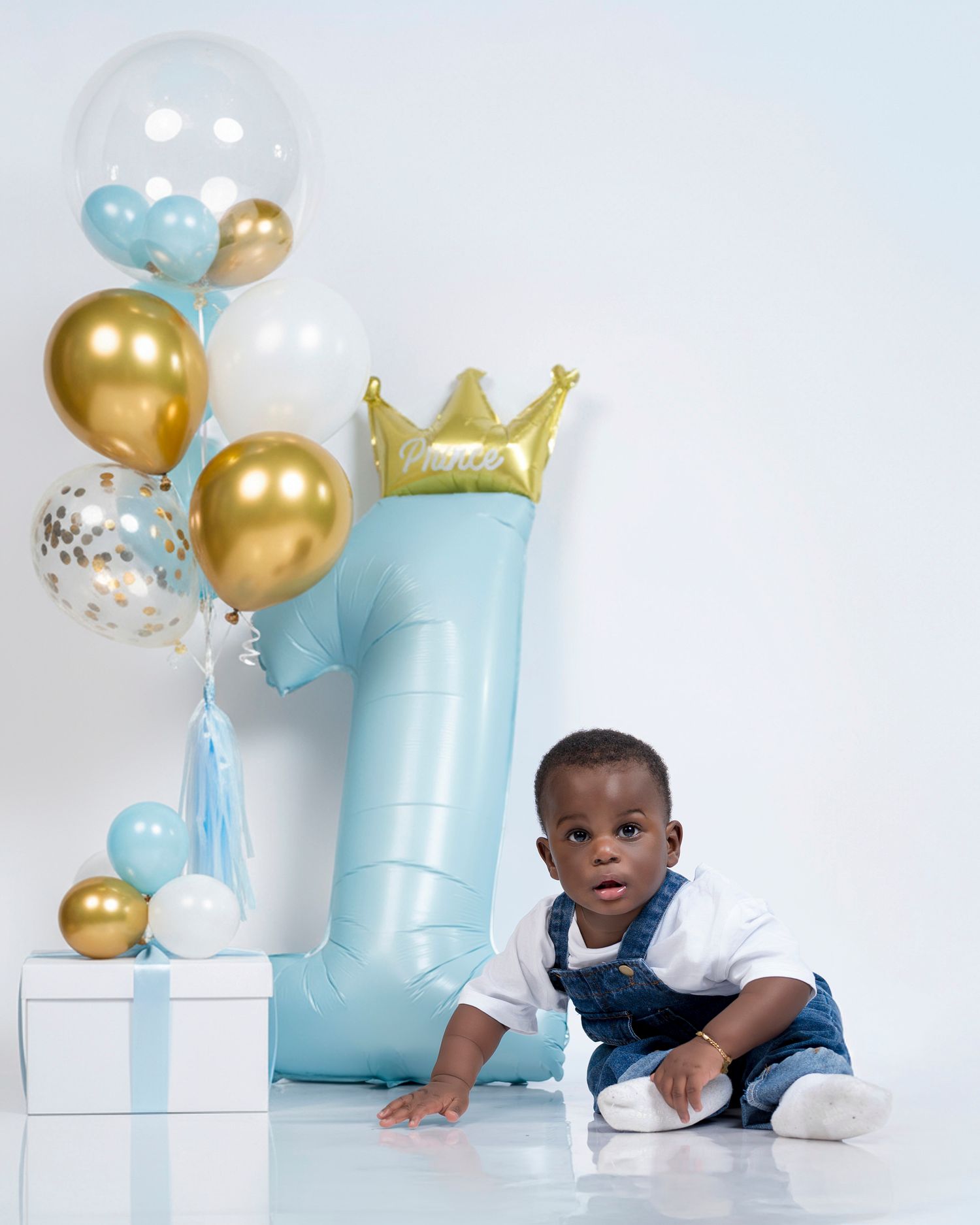 Baby in white and navy outfit sits among blue and gold balloon decorations for first birthday celebration.