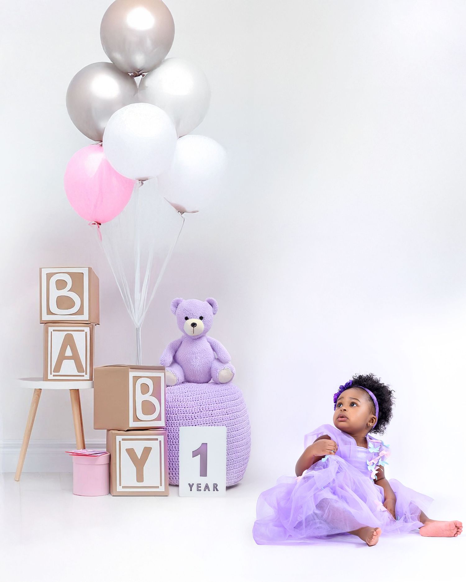 First birthday photo setup with purple decor featuring white and silver balloons, alphabet blocks, and purple teddy bear.