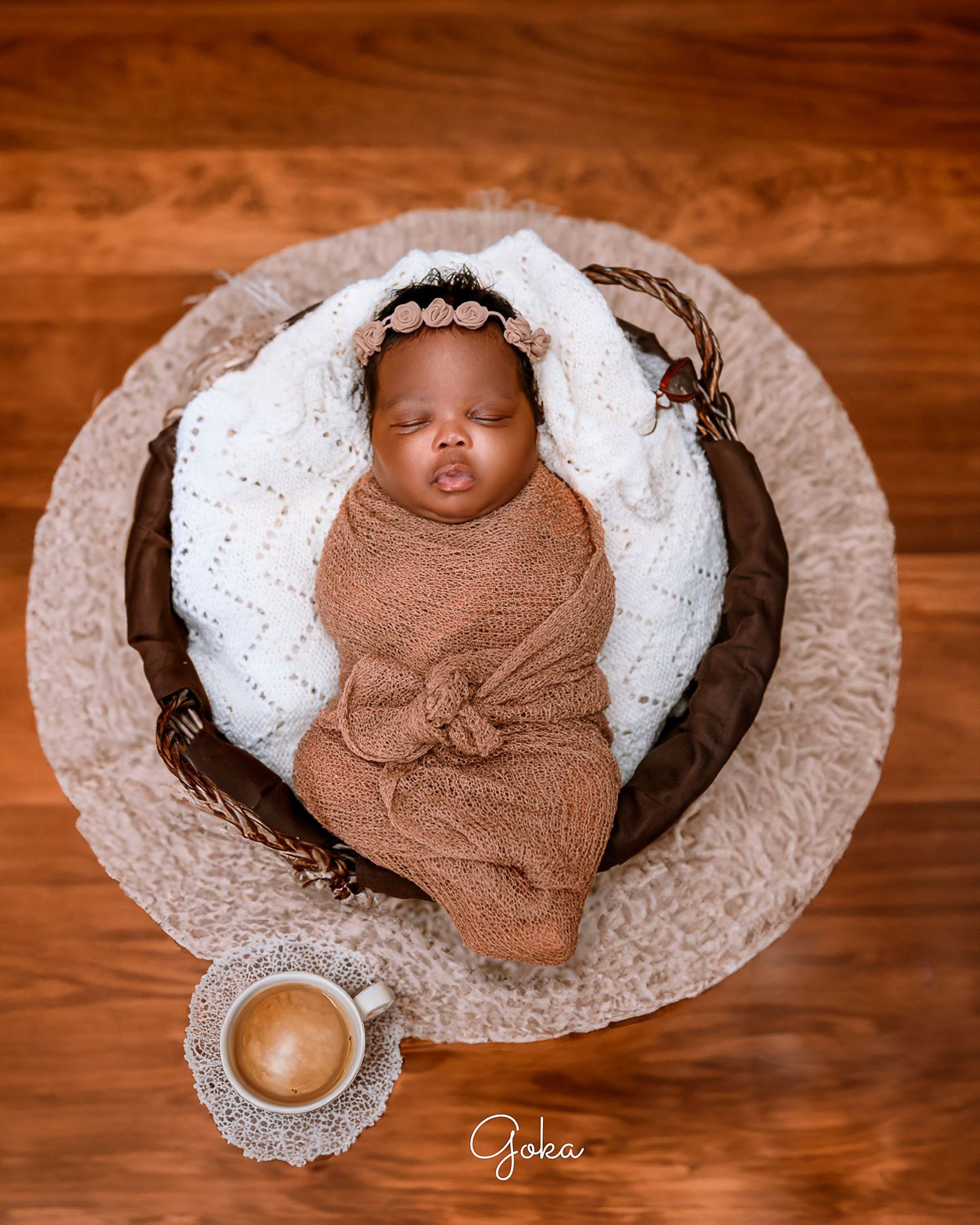 Sleeping baby wrapped in brown knit blanket nestled in white cushioned basket with coffee cup decor.