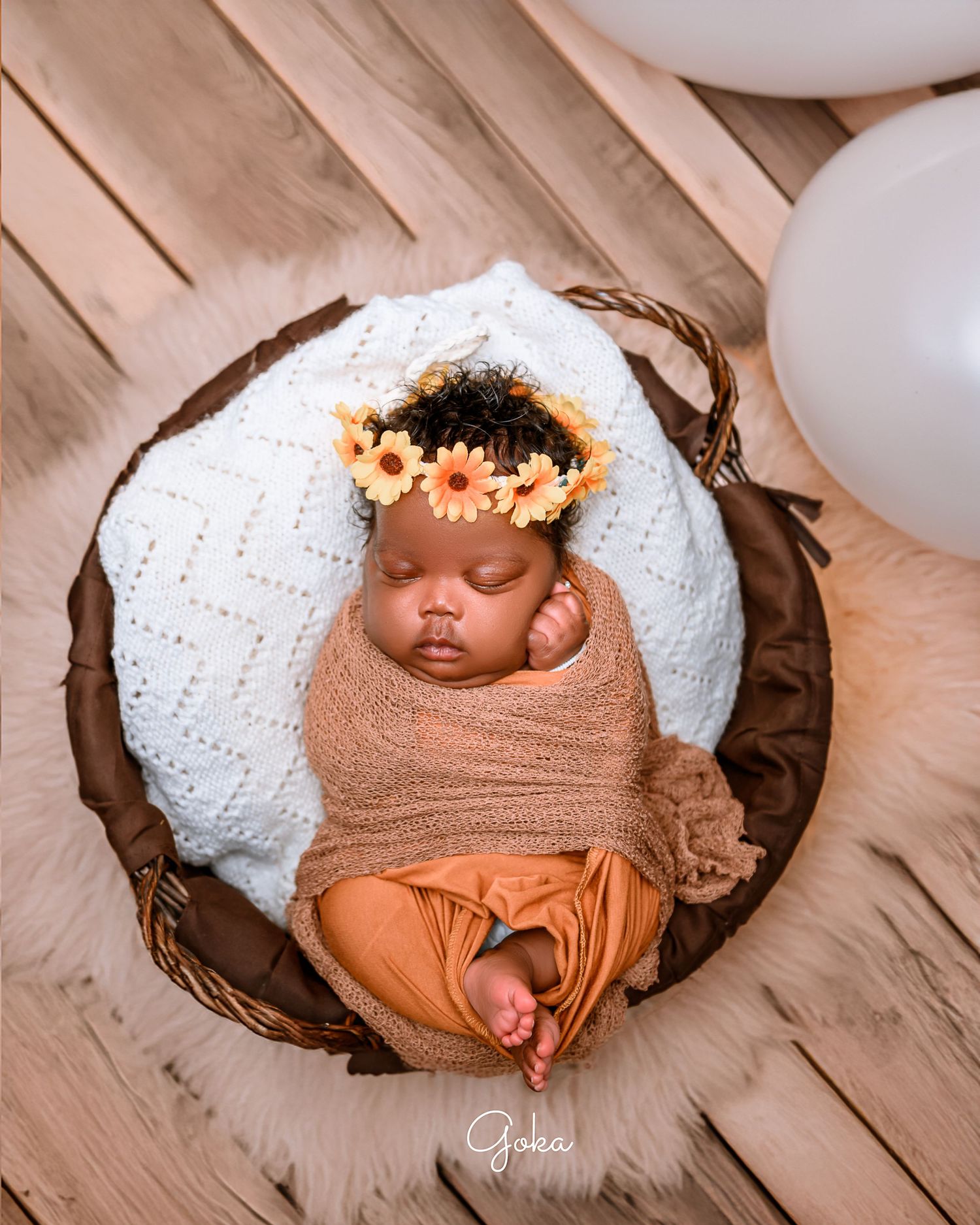 Sleeping newborn in brown wrap with floral crown in white cushioned basket.