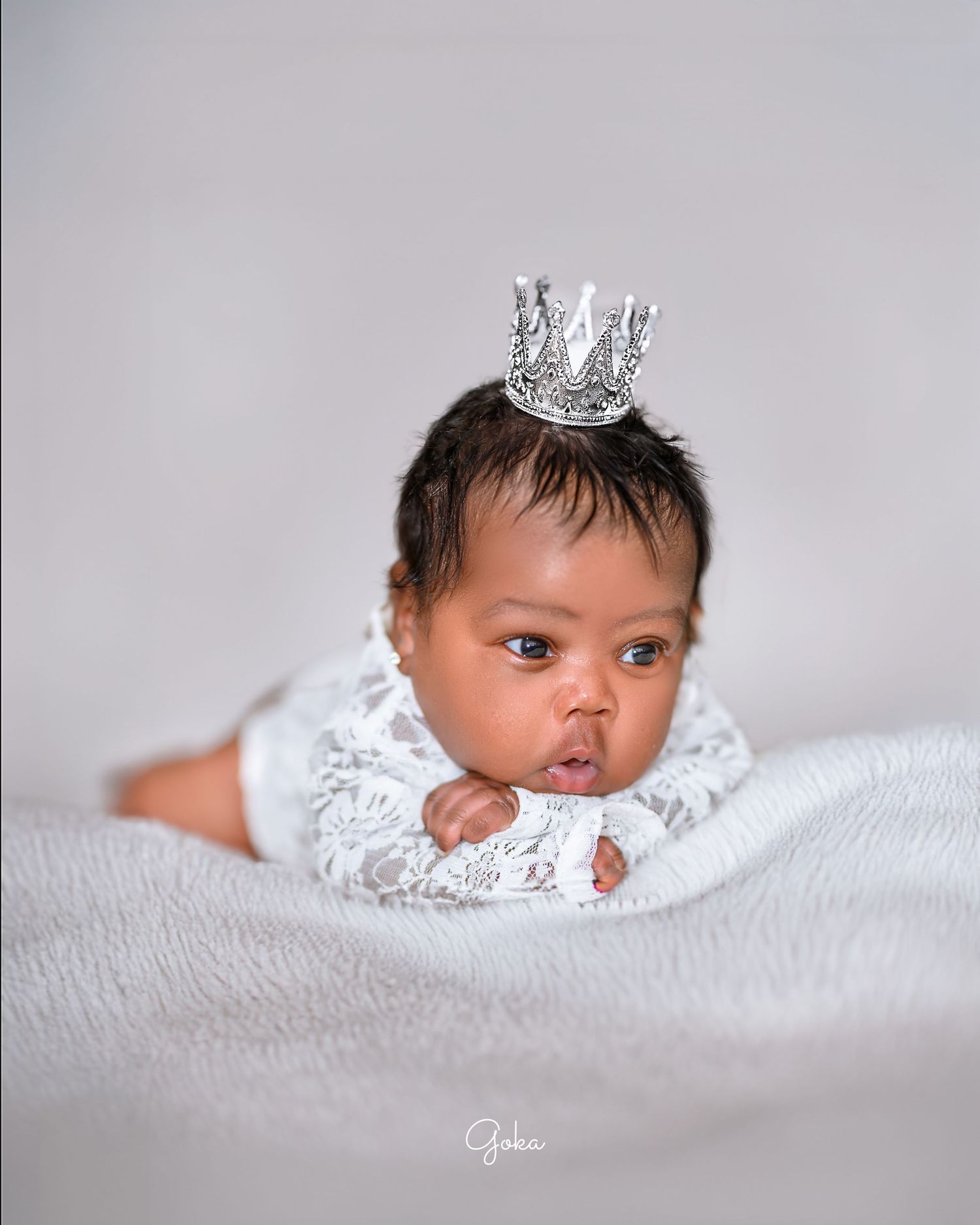Baby wearing tiny silver crown and lace outfit lying on white bedding.