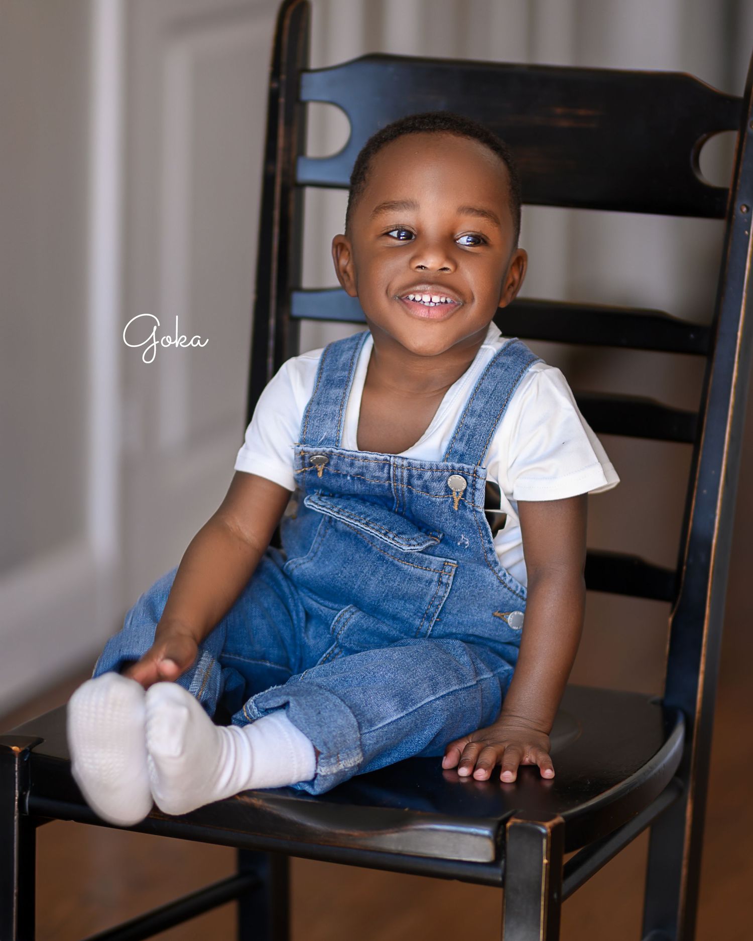 Portrait of toddler in denim overalls sitting on wooden chair and smiling.