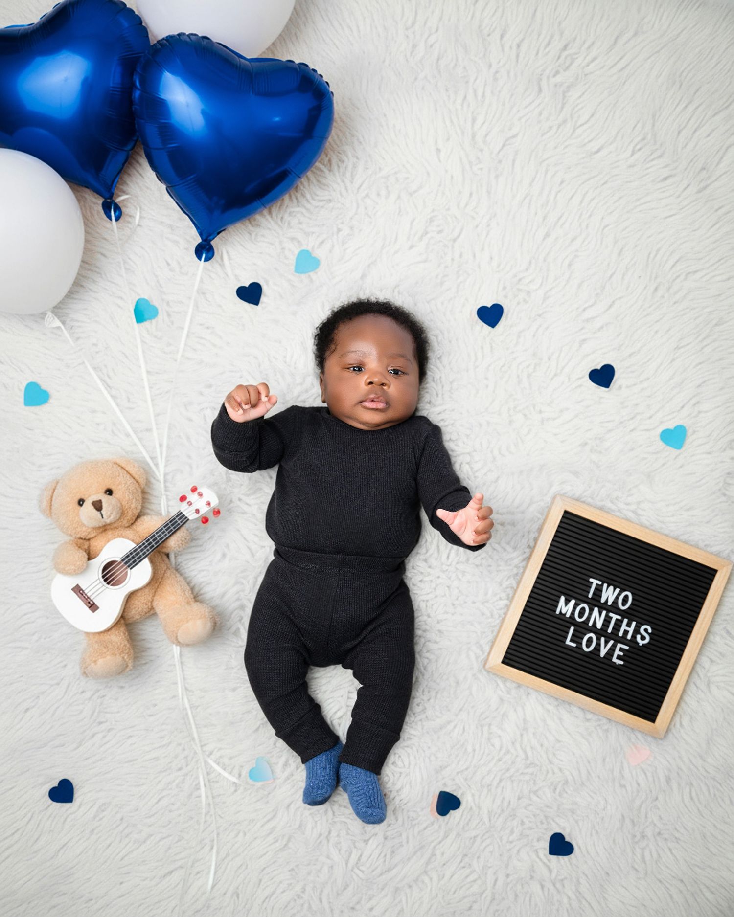 Baby in black outfit lying on white blanket with blue heart balloons and teddy bear for milestone photoshoot.