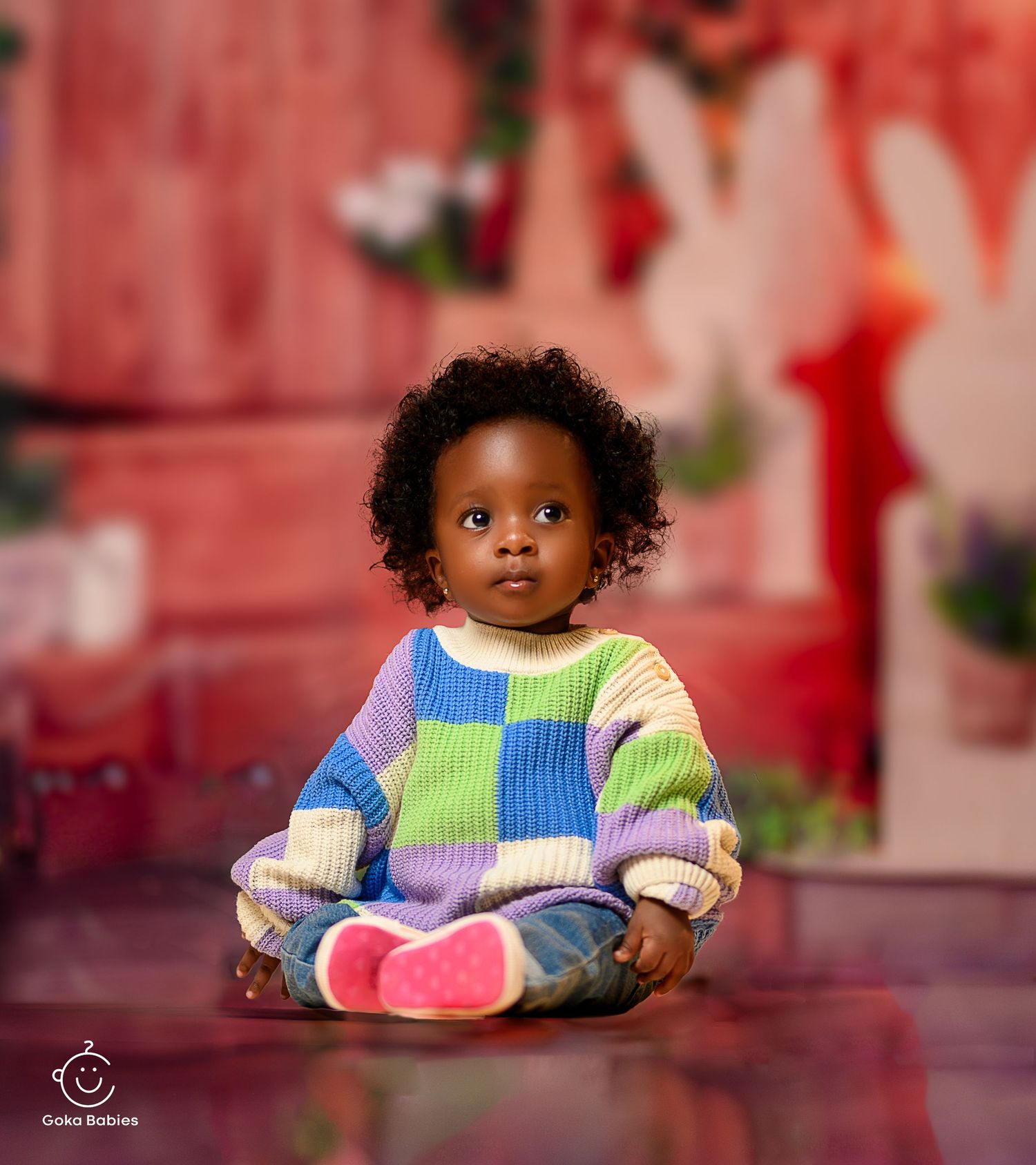 Baby in colorful patchwork sweater sitting cross-legged on red studio background.