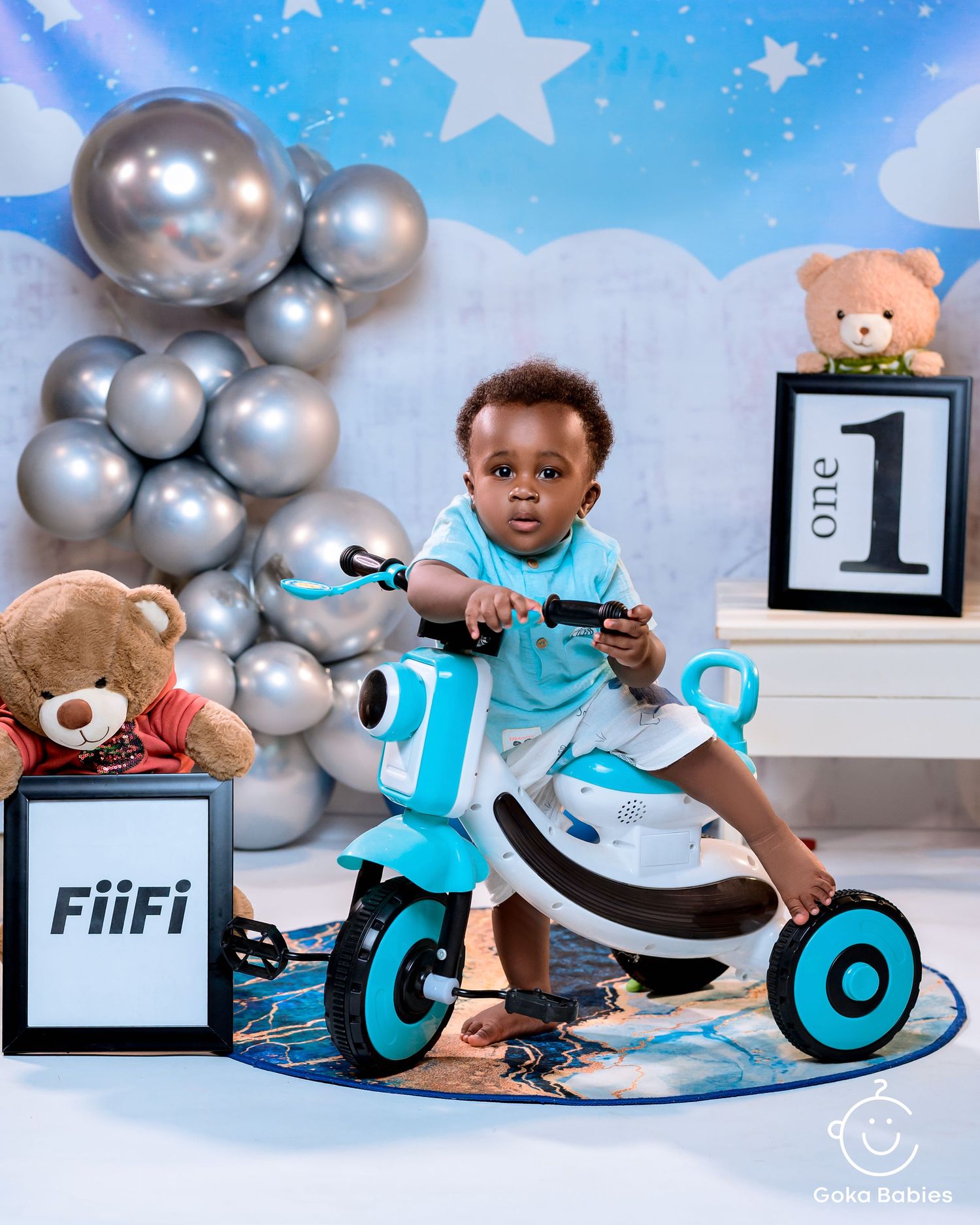 Child riding turquoise tricycle against starry backdrop with silver balloons.
