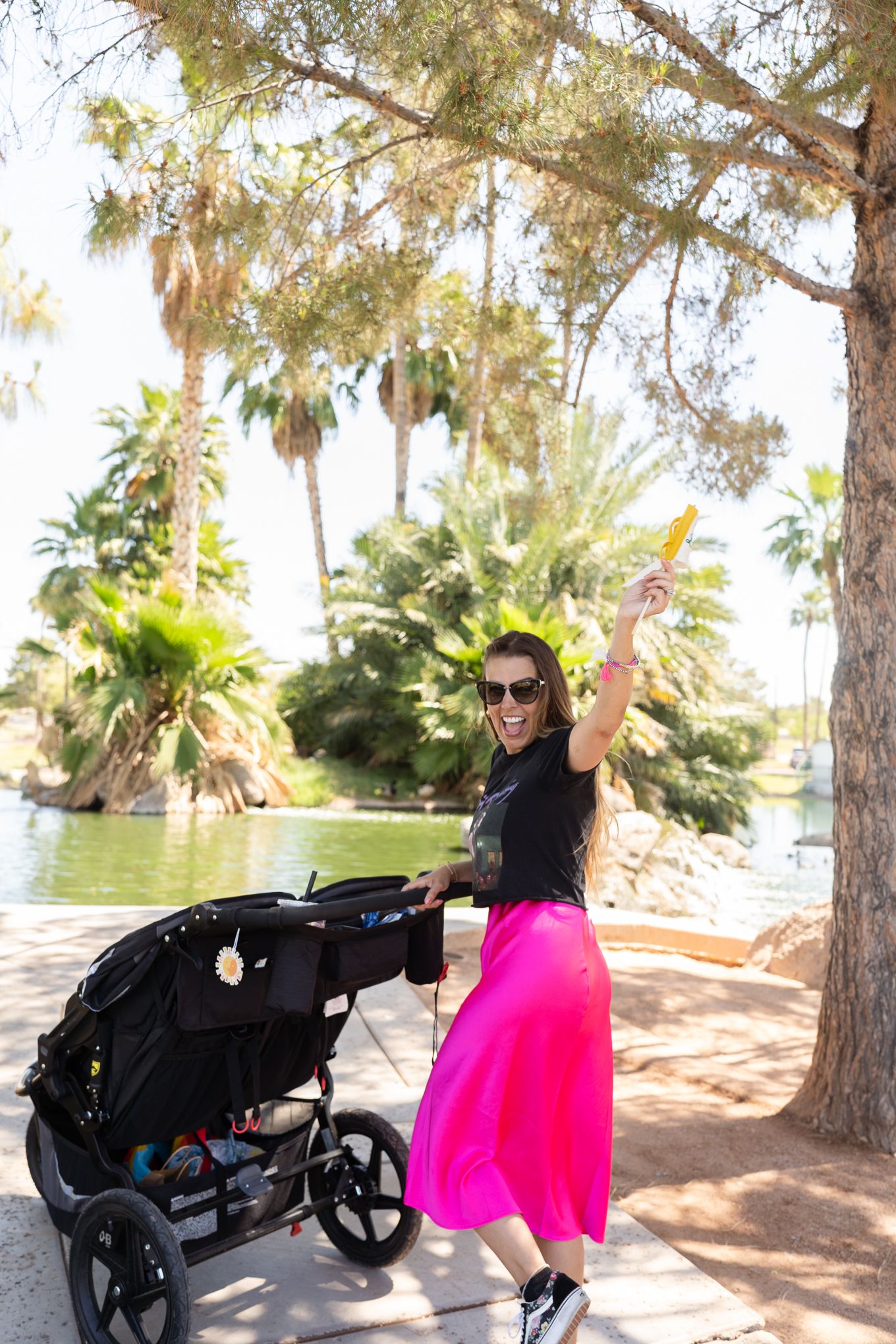 A person wearing a bright pink skirt poses joyfully near palm trees and a scenic pond.