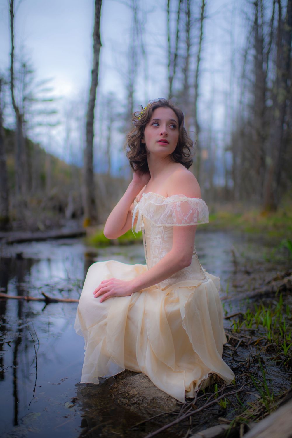 Woman in Victorian-style dress kneels in shallow swamp water surrounded by bare winter trees.