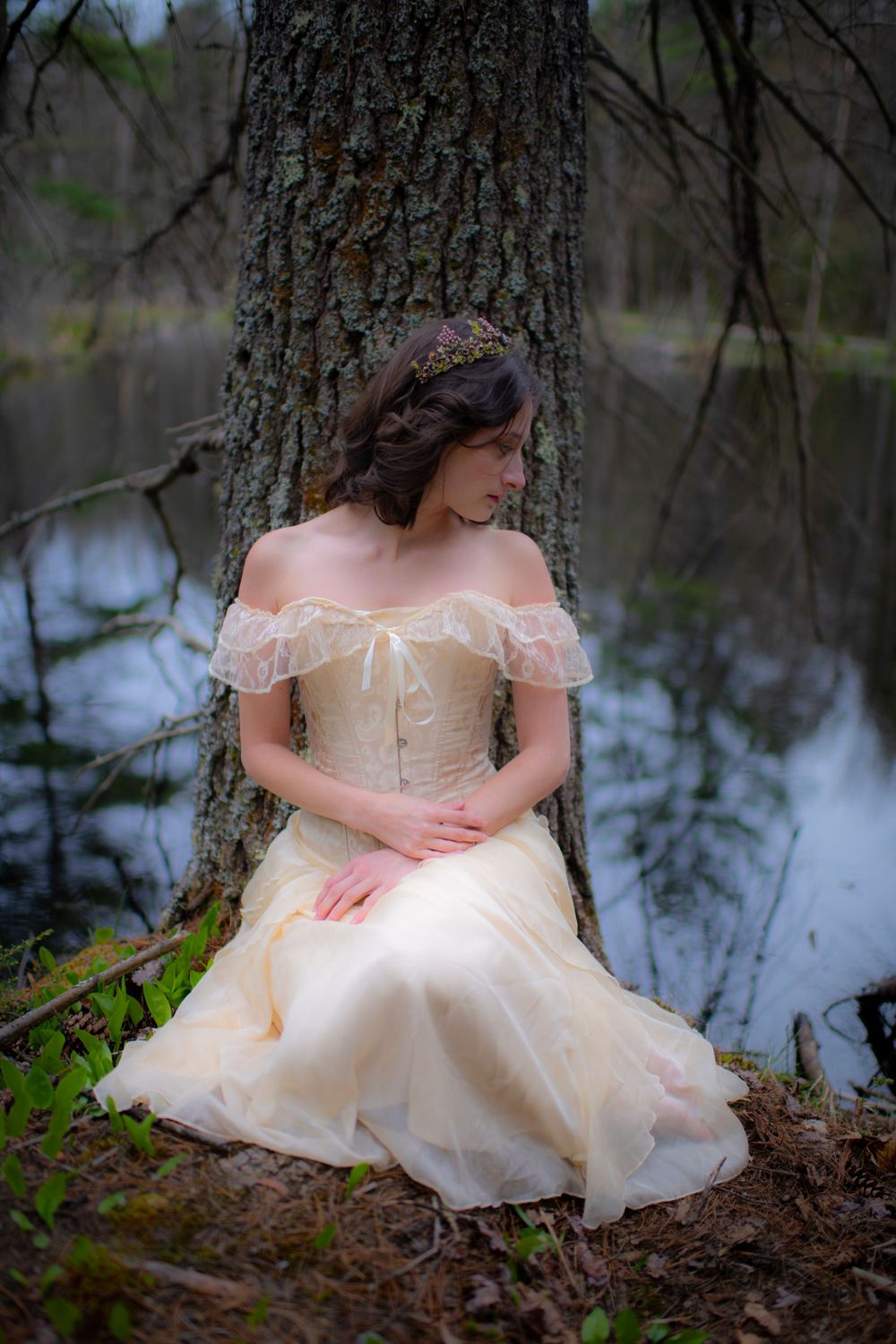 Woman in delicate off-shoulder dress poses against tree trunk near calm water.