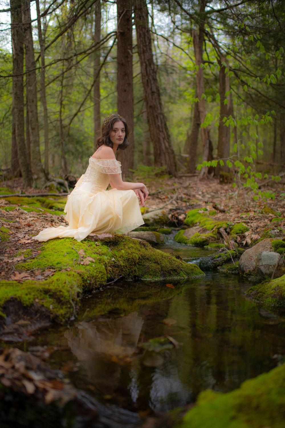 Romantic woodland photograph by a reflective stream featuring cream-colored vintage dress and mossy stones.