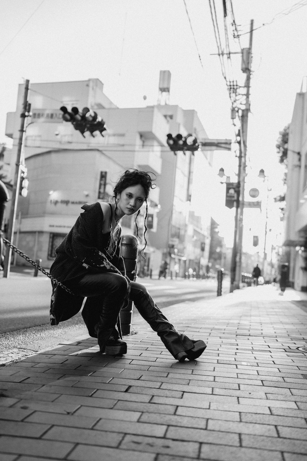 Black and white street photography of a woman resting by the road against the chain fence