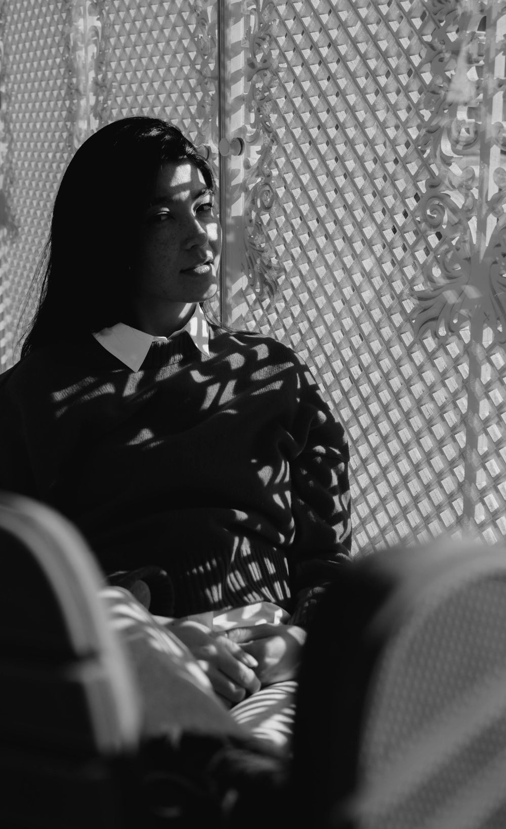 Black and white portrait of a woman sitting by a patterned wall with sunlight casting intricate patterns on her